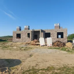 Budowa domu jednorodzinnego w Wilkowie w trakcie wznoszenia ścian z bloczków betonowych. Widoczne materiały budowlane, rusztowanie i niezabudowane otwory okienne.