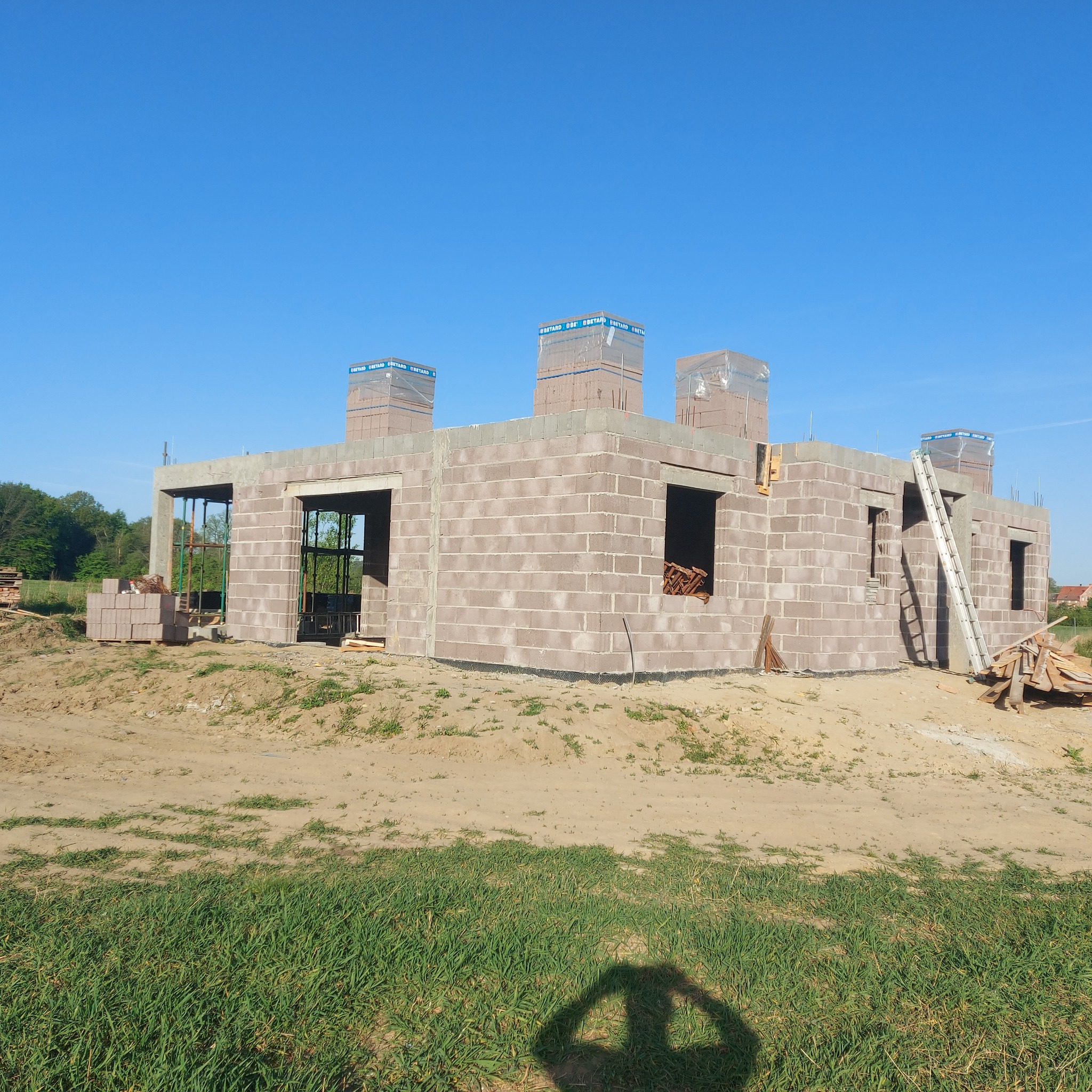 Surowy stan domu z betonu komórkowego, okna zabite deskami, częściowo otynkowane kominy zabezpieczone folią, drabina oparta o ścianę, widoczne materiały budowlane.