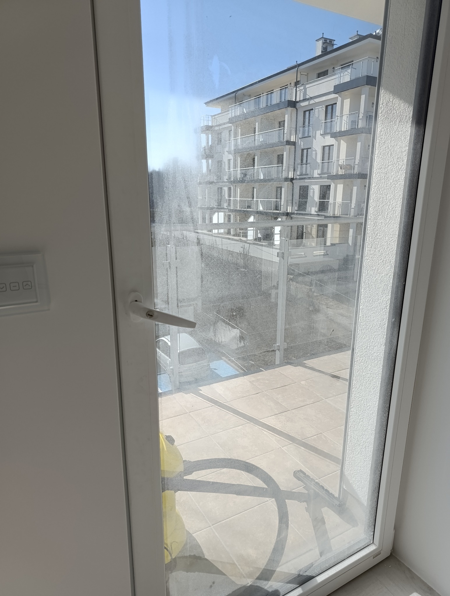Widok przez brudne okno na budynek z balkonami. Na pierwszym planie narzędzia do mycia okien, sugerujące trwający proces czyszczenia.