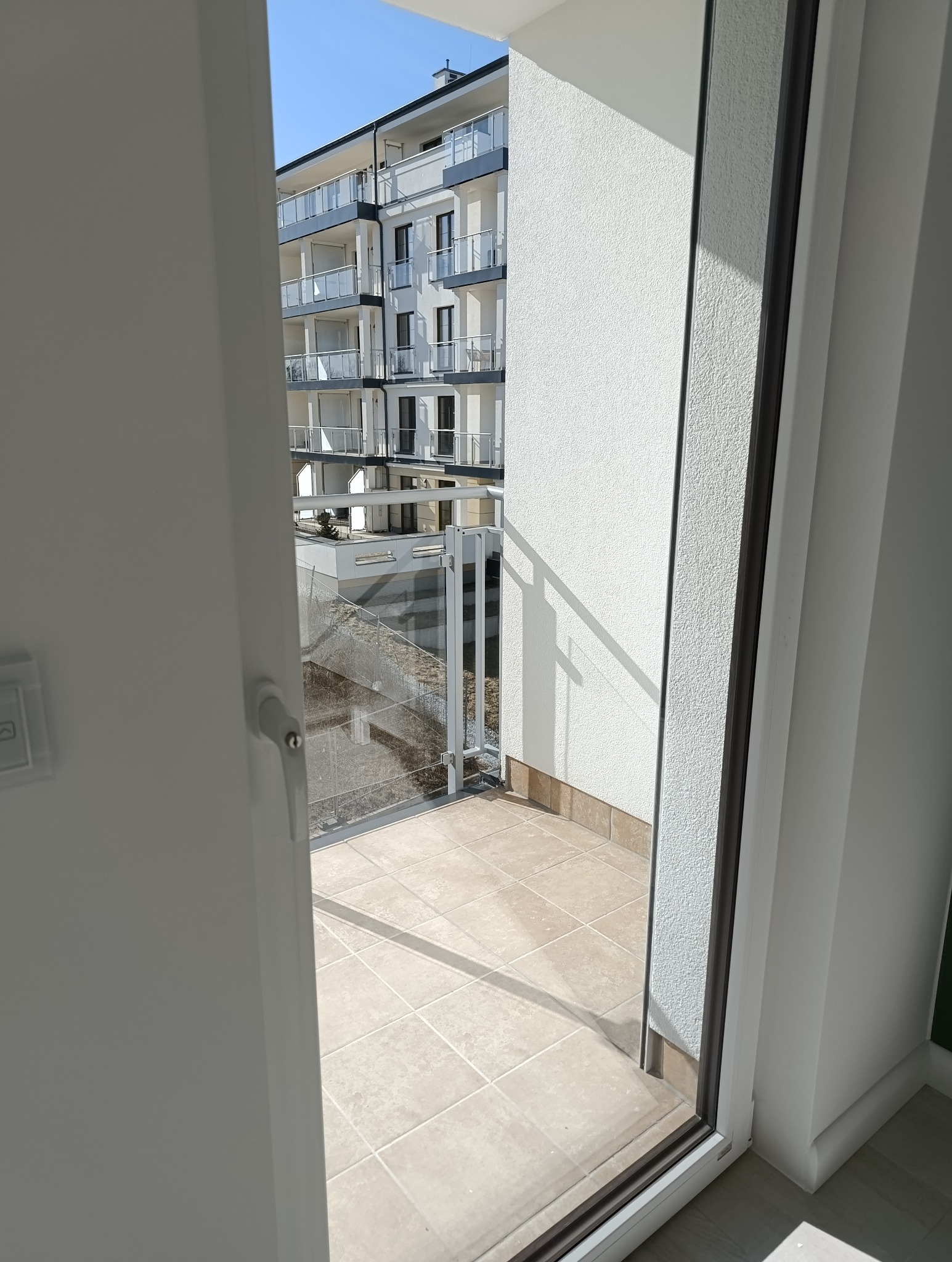 Widok z wnętrza na czysty, jasny balkon z beżowymi płytkami i szklaną balustradą. W tle budynek mieszkalny z balkonami. Perspektywa zza otwartych drzwi balkonowych.