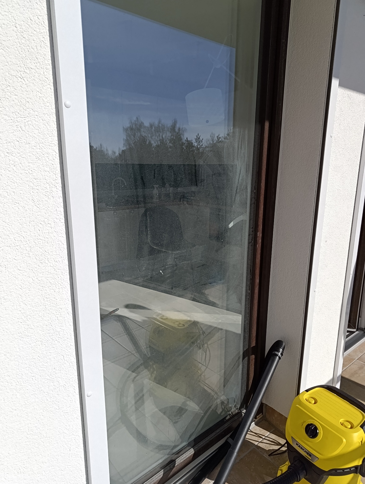 Brudna szyba okienna z widocznym odkurzaczem Kärcher. Widoczne zacieki i kurz na szkle. Biała rama okienna i fragment elewacji budynku.