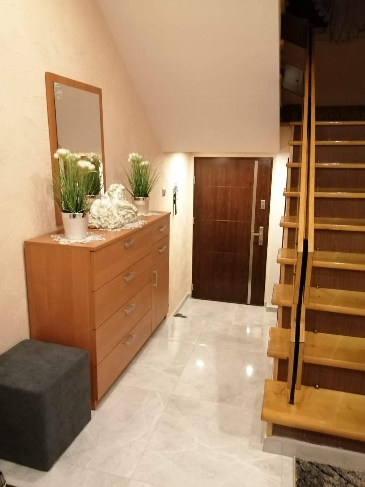 Jasne wnętrze przedpokoju z drewnianą komodą, lustrem, kwiatami w doniczkach, rzeźbą, szarym pufem, drewnianymi schodami i ciemnymi drzwiami.