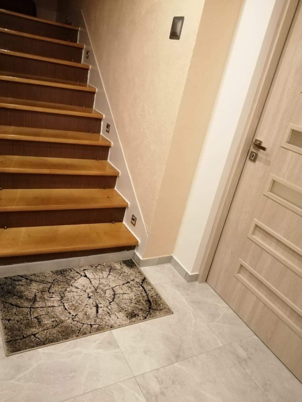 Drewniane schody z oświetleniem punktowym na ścianie, szary dywanik imitujący przekrój pnia drzewa, jasne płytki podłogowe i drzwi z przeszkleniami.