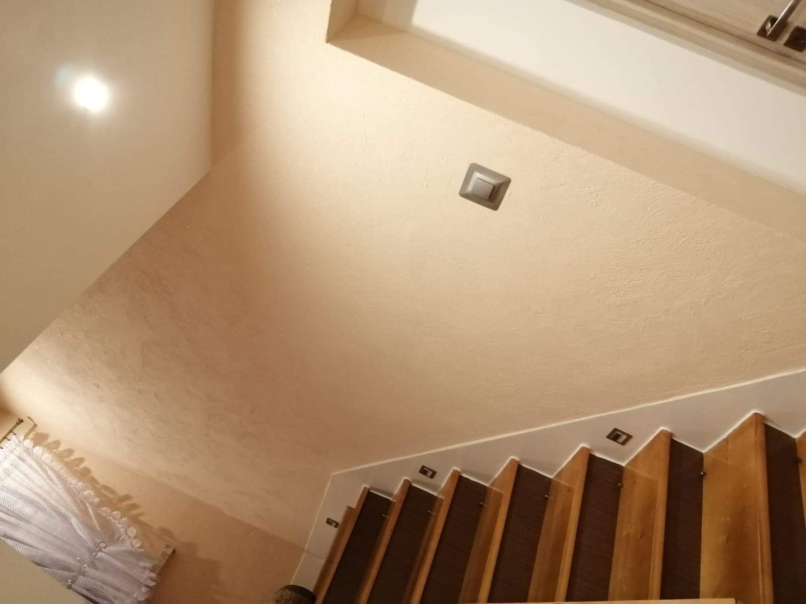 Beżowa ściana i sufit z wbudowanym oświetleniem schodów, widok z dołu na drewniane schody z ciemnymi panelami, fragment zasłony w rogu.