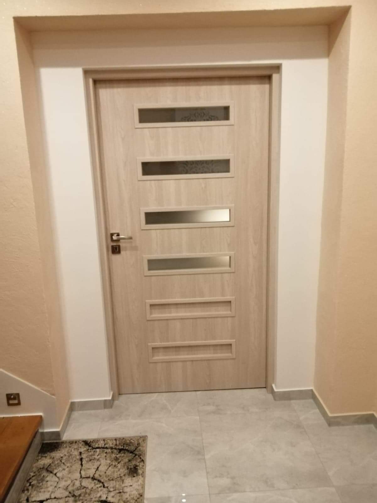 Jasna, drewnopodobna wewnętrzna drzwi z czterema podłużnymi przeszkleniami, zamontowana w białej ościeżnicy w beżowym korytarzu z szarą podłogą.