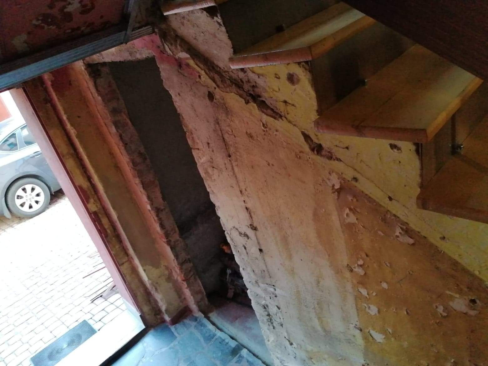 Schody drewniane w trakcie remontu, widoczne ślady starej farby i uszkodzenia na ścianie, z boku zaparkowany samochód.