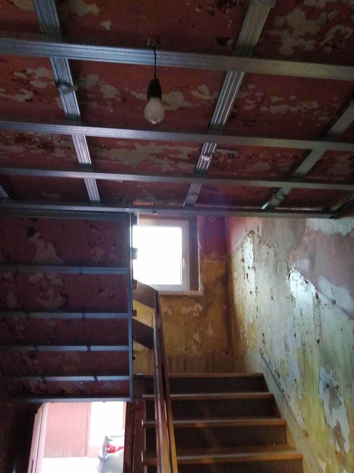Stare wnętrze w trakcie prac: widok na drewniane schody, okno, ściany z odpadającą farbą i konstrukcję sufitu podwieszanego z metalowych profili oraz wiszącą żarówkę.