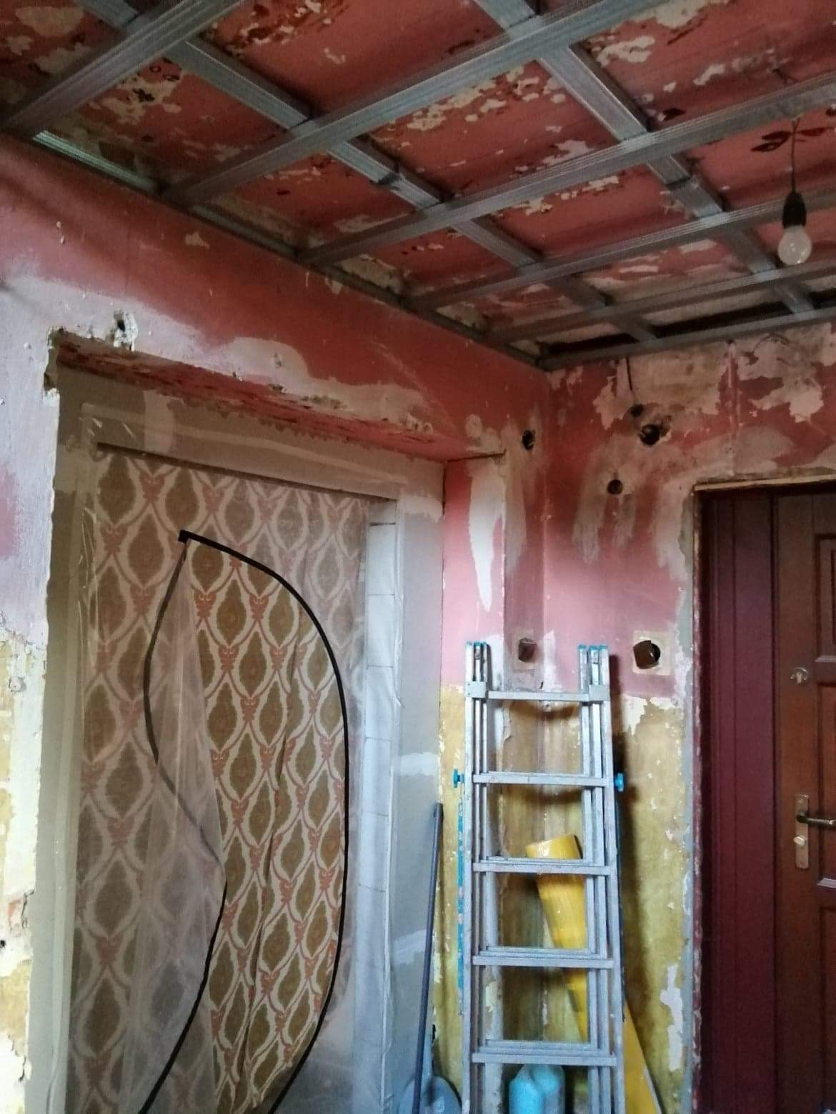 Pokój w trakcie prac przygotowawczych: zerwane tapety, odsłonięte instalacje elektryczne, metalowa konstrukcja sufitu podwieszanego, drabina aluminiowa z żółtym wiadrem i niebieskim płynem...