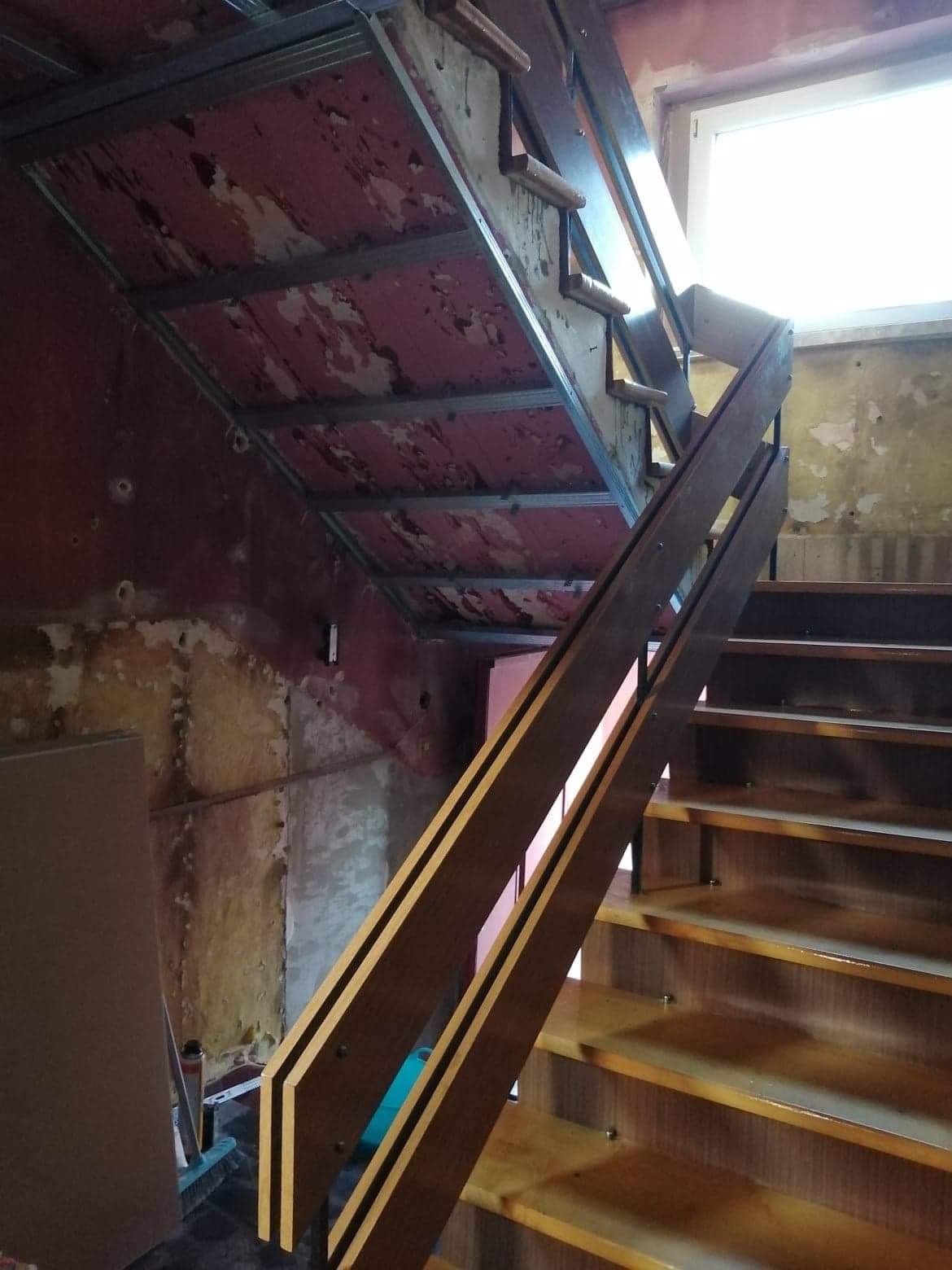 Klatka schodowa w trakcie prac wykończeniowych: drewniane schody z barierką, surowy sufit z metalową konstrukcją i ściany z widocznymi śladami po farbie.