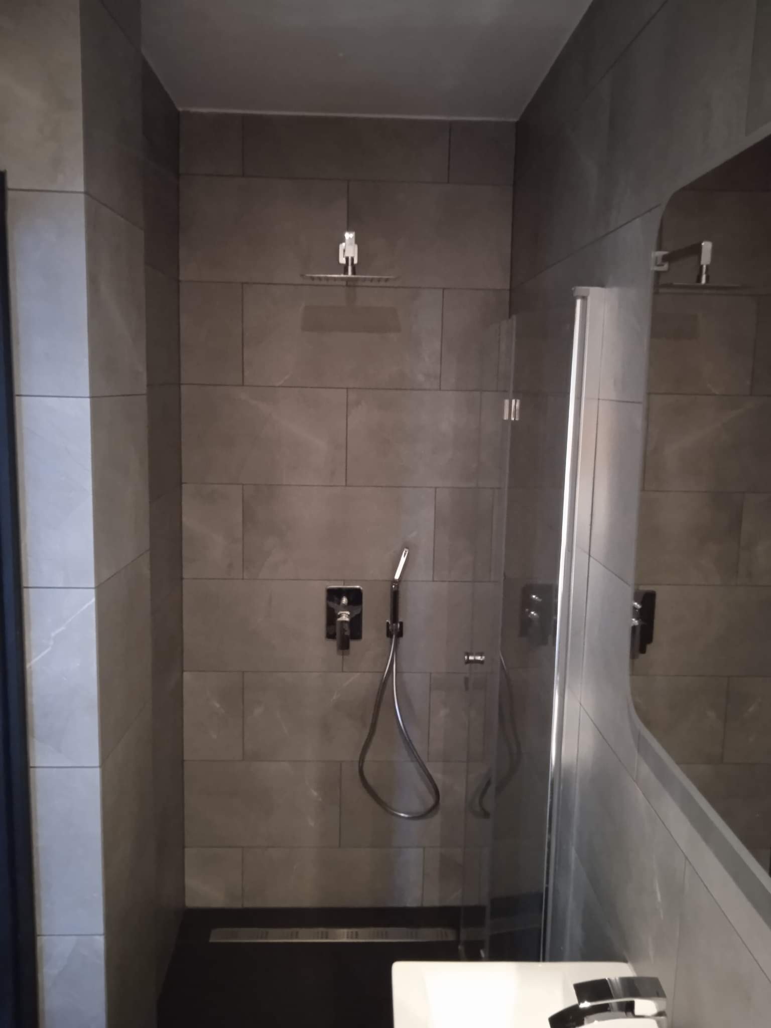 Nowoczesna kabina prysznicowa z szarymi płytkami imitującymi kamień, czarną armaturą i odpływem liniowym.