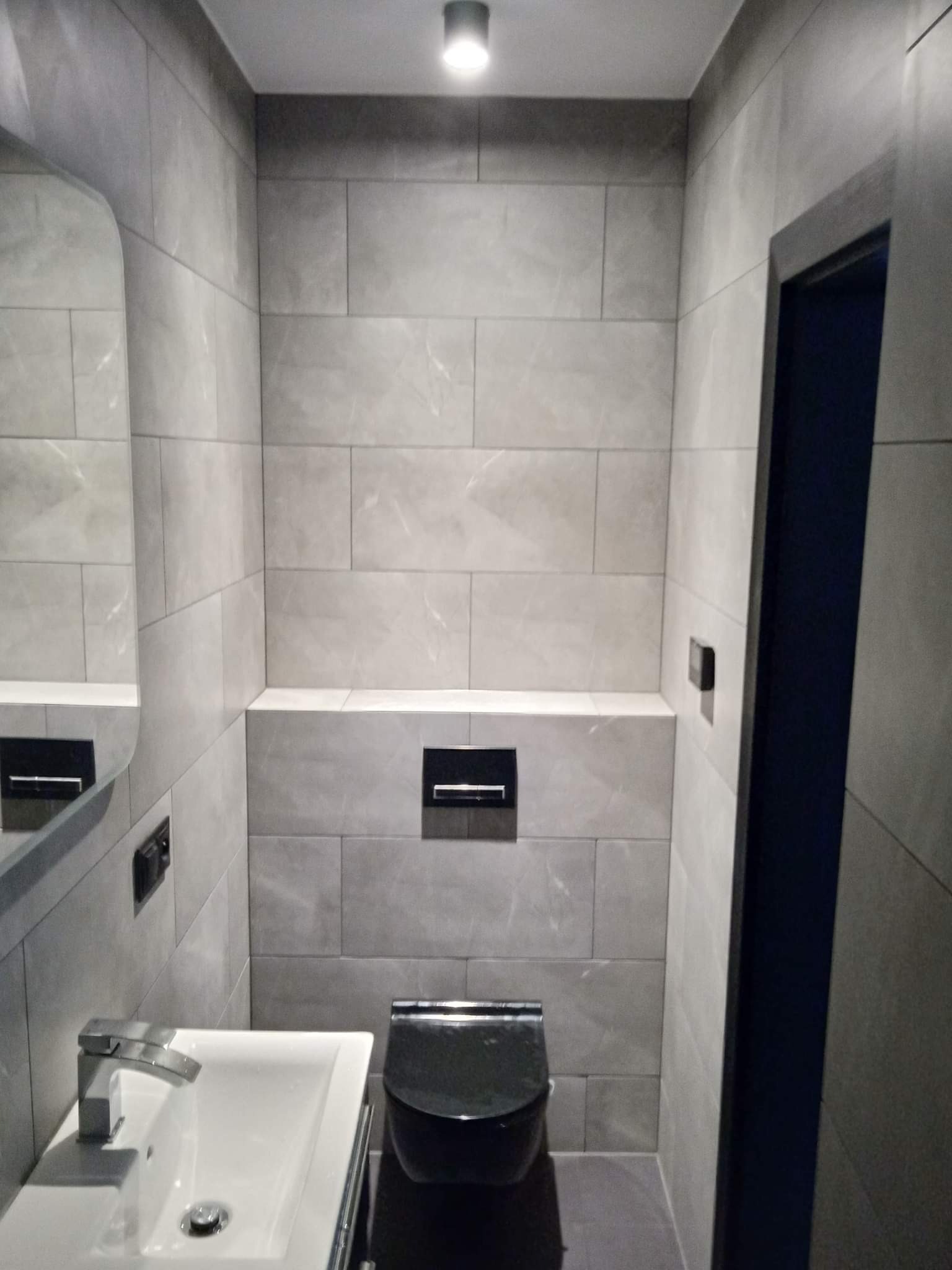 Nowoczesna, szara łazienka w Kornowacu z podwieszaną toaletą, umywalką z chromowaną baterią i lustrem bez ramy, wykończona płytkami imitującymi kamień.