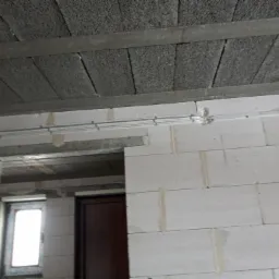 Surowe wnętrze w trakcie budowy z widocznymi przewodami elektrycznymi prowadzonymi po ścianie z bloczków betonowych i stropem z płyt betonowych.