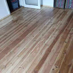 Jasne, lakierowane deski podłogowe z widocznym usłojeniem, ułożone ukośnie w pomieszczeniu z białymi ścianami i fragmentami drzwi oraz dekoracyjnych paneli.