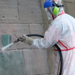 Pracownik w kombinezonie ochronnym i z osłoną twarzy czyści kamienną ścianę strumieniem wody pod ciśnieniem, widoczne różnice w kolorze pomiędzy oczyszczonym a nieoczyszczonym fragmentem.