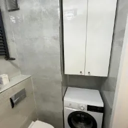 Szara łazienka z pralką Electrolux, szafką wiszącą i podtynkową toaletą. Minimalistyczny design z płytkami imitującymi beton. Nowoczesne wykończenie wnętrza.