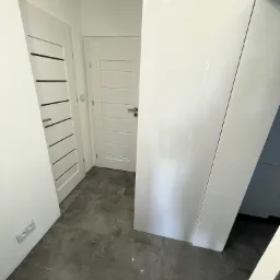 Nowoczesny przedpokój z dwiema białymi parami drzwi o różnych wzorach i szarą podłogą z płytek. Widoczne gniazdko elektryczne.