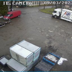 DBK Logistics - Widok z kamery IP na plac magazynowy z kontenerami, wózkiem widłowym i ciężarówką z niebieskimi kontenerami, w tle czerwony bus i fragment ramy przyczepy