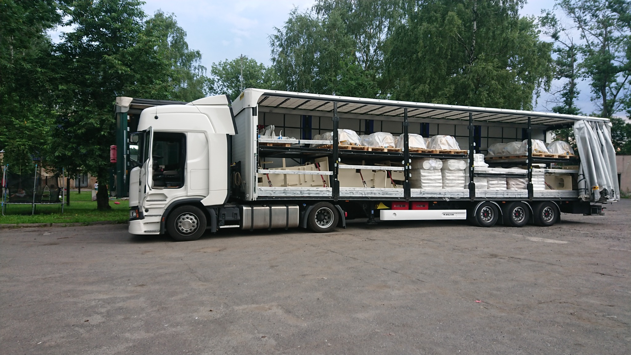 Otwarta naczepa ciężarówki z wielopoziomowym systemem załadunku, przewożąca zabezpieczone folią palety z elementami wyposażenia łazienek i ceramiką sanitarną.