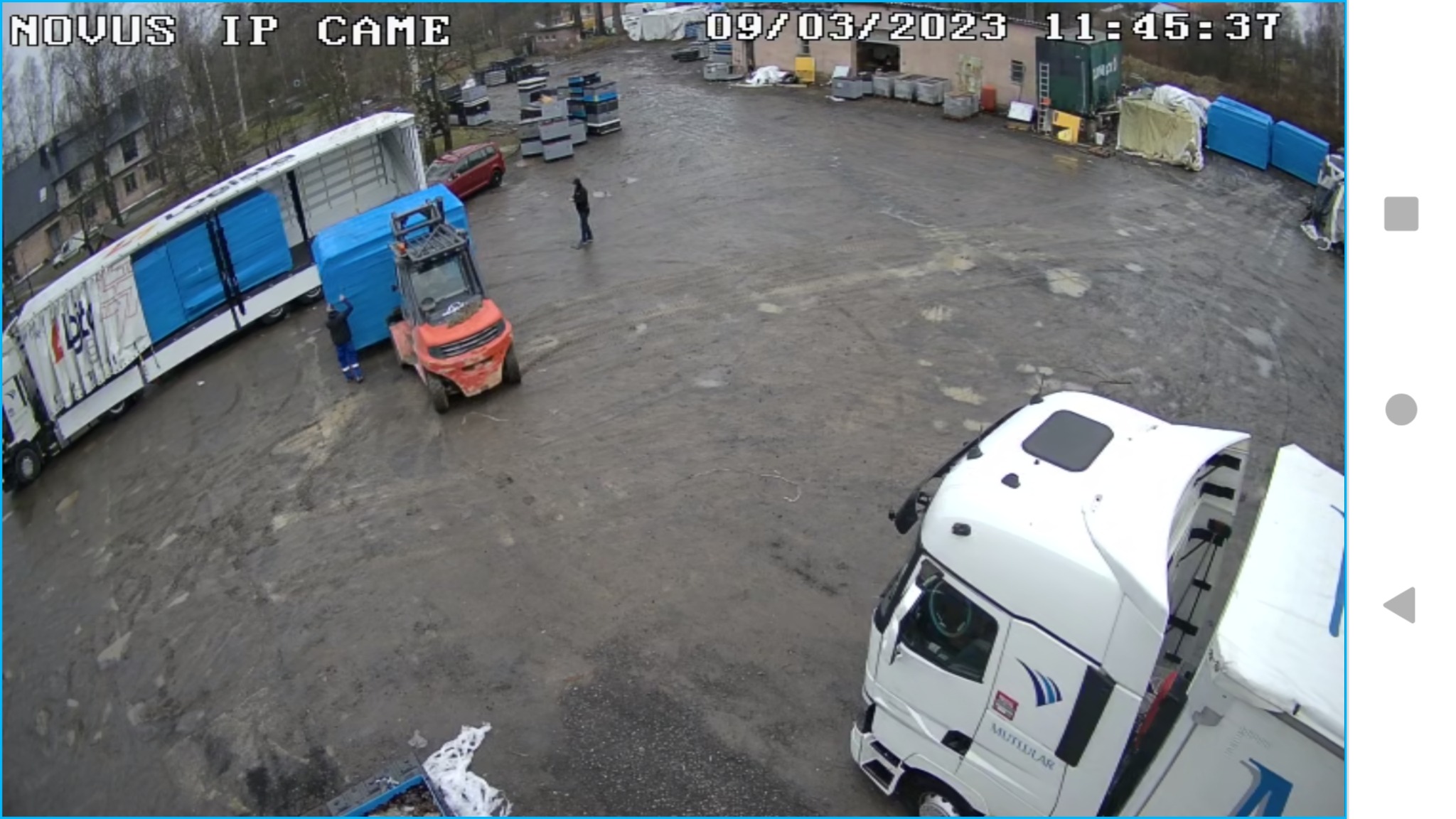 Ujęcie z kamery przemysłowej w Pieńsku przedstawia załadunek niebieskich elementów konstrukcyjnych na naczepę ciężarówki za pomocą wózka widłowego. W tle widoczny plac z materiałami budowlanymi.