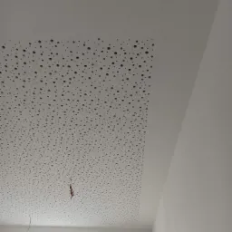 Biały sufit akustyczny z regularnymi otworami, częściowo zamontowany, z widocznymi przewodami i oknem w tle.