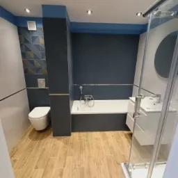 Nowoczesna łazienka po remoncie z drewnopodobną podłogą, ciemną ścianą z glazury, białą armaturą i kabiną prysznicową z przezroczystego szkła.