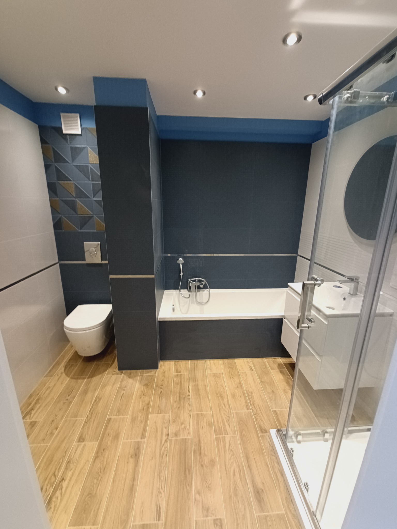 Nowoczesna łazienka po remoncie z drewnopodobną podłogą, ciemną ścianą z glazury, białą armaturą i kabiną prysznicową z przezroczystego szkła.