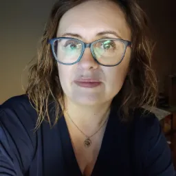 Portret kobiety w okularach, z delikatnym uśmiechem, w ciemnym ubraniu i ze złotym naszyjnikiem, w słabym oświetleniu.