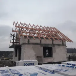 Surowy stan domu z pustaków Solbet z widoczną konstrukcją więźby dachowej, palety z materiałem budowlanym na pierwszym planie, pochmurne niebo w tle.