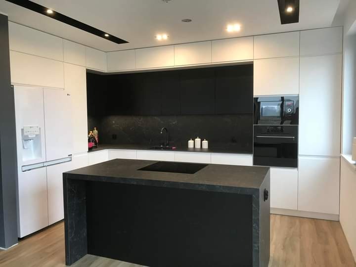 Nowoczesna kuchnia z wyspą, białe szafki górne i dolne kontrastują z czarnymi elementami, takimi jak blat wyspy i ściana nad blatem, oświetlenie punktowe w suficie.