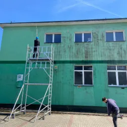 Dwóch mężczyzn przy zielonej elewacji budynku. Jeden na rusztowaniu, drugi na ziemi. Widoczne ślady zabrudzeń na ścianie, sugerujące proces czyszczenia.