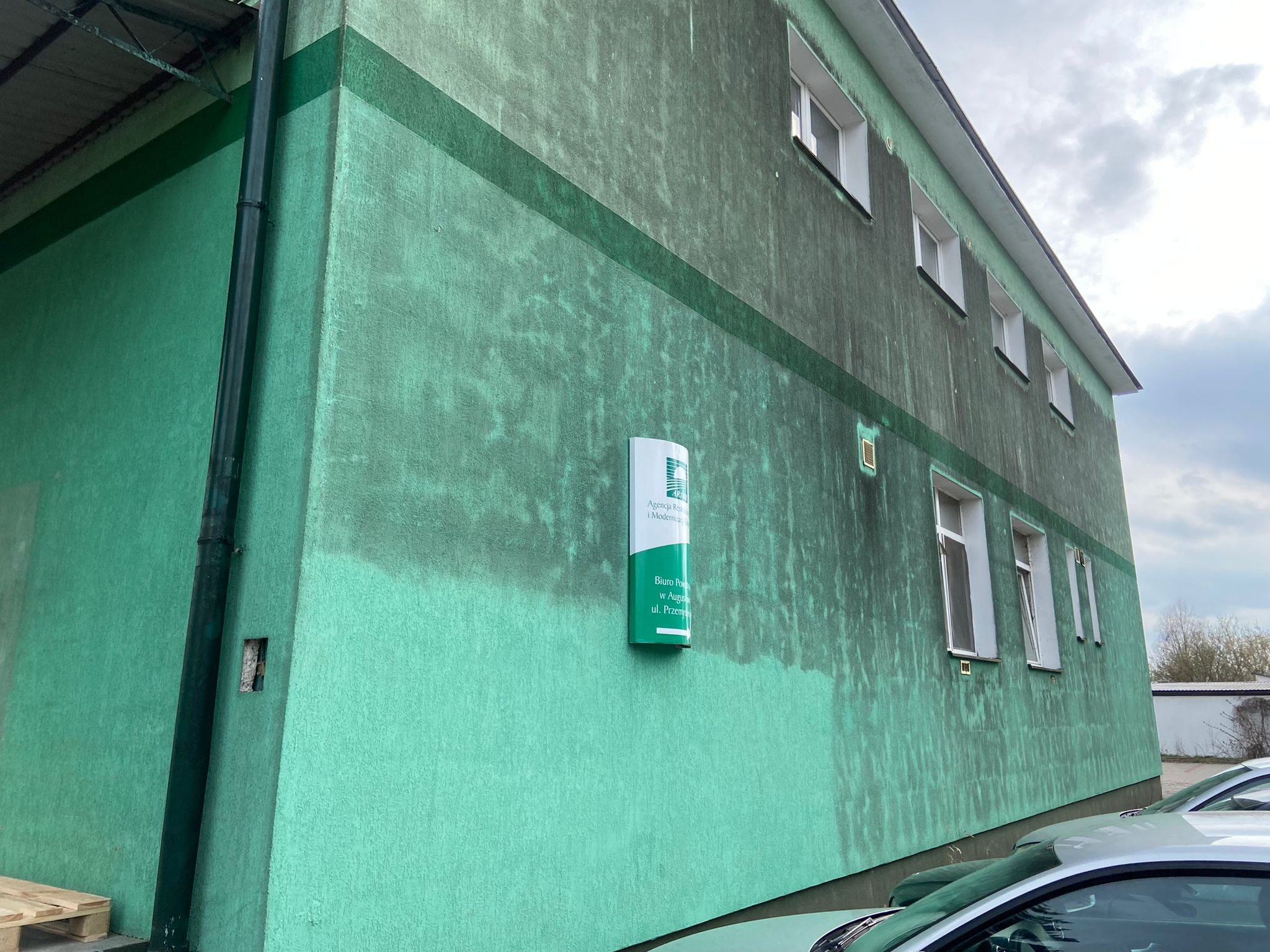 Zielona elewacja budynku z widocznymi śladami zabrudzeń i zacieków, sugerująca potrzebę odświeżenia lub umycia. Widoczny fragment rynny i zaparkowane auta.