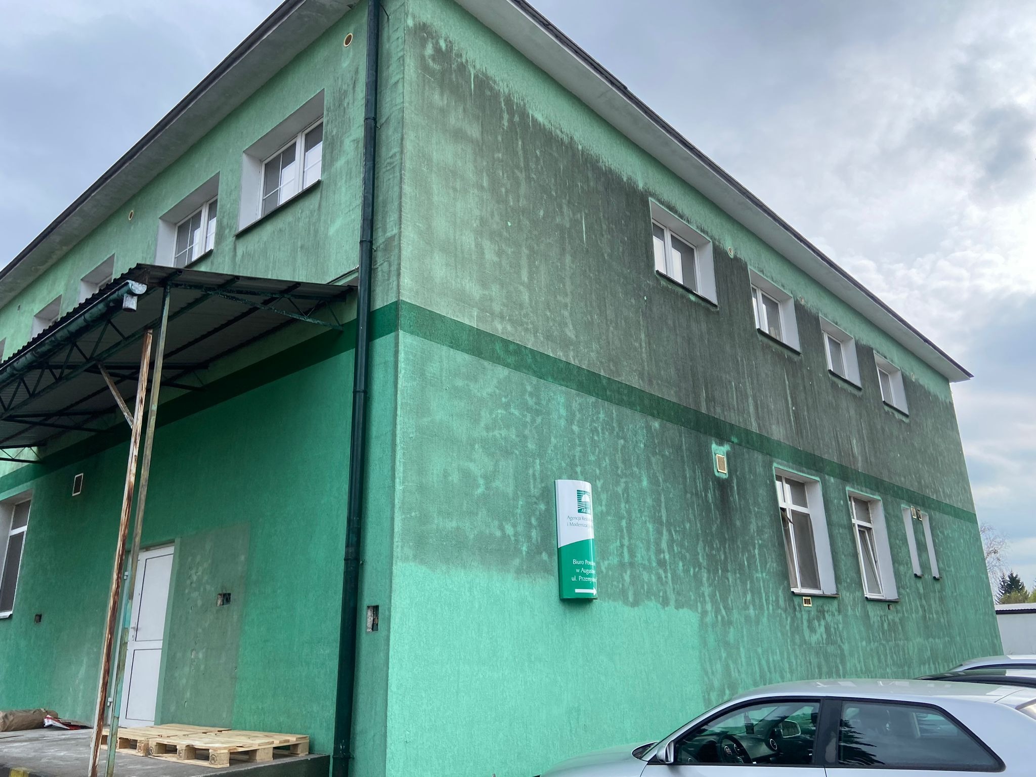 Budynek biurowy z widoczną różnicą w czystości elewacji, sugerującą częściowe umycie. Fragment zaparkowanego auta w prawym dolnym rogu.