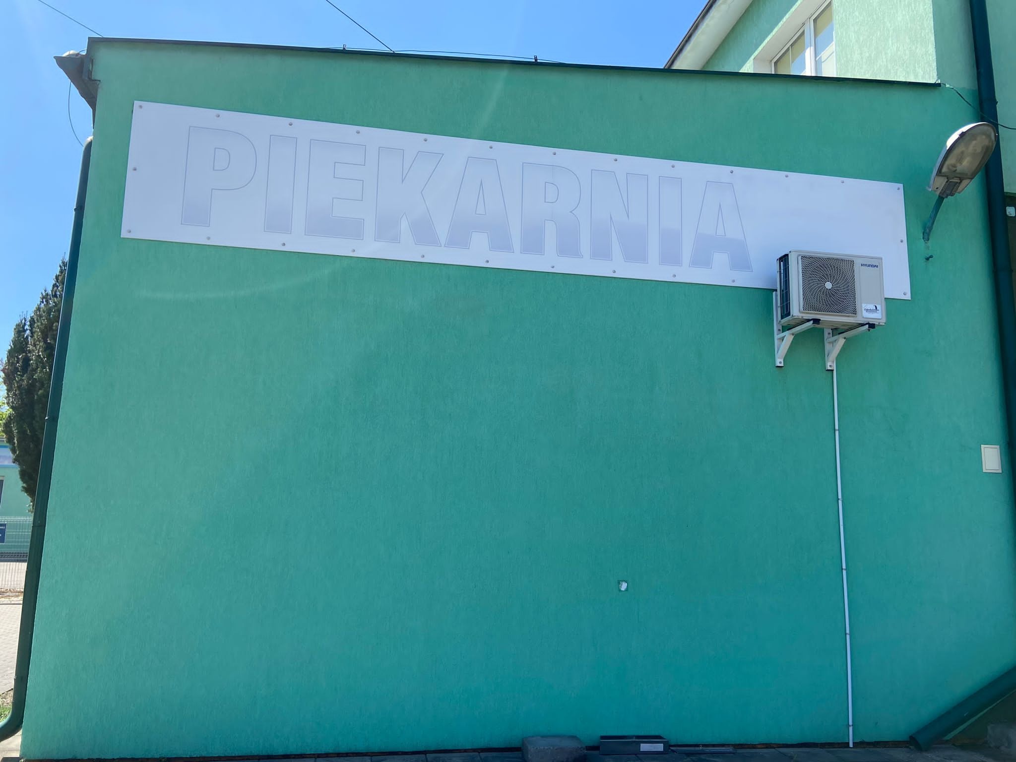 Turkusowa elewacja budynku z szyldem 'Piekarnia', klimatyzatorem i lampą uliczną. Świeża, jednolita farba. Widoczne detale konstrukcyjne i elementy otoczenia.