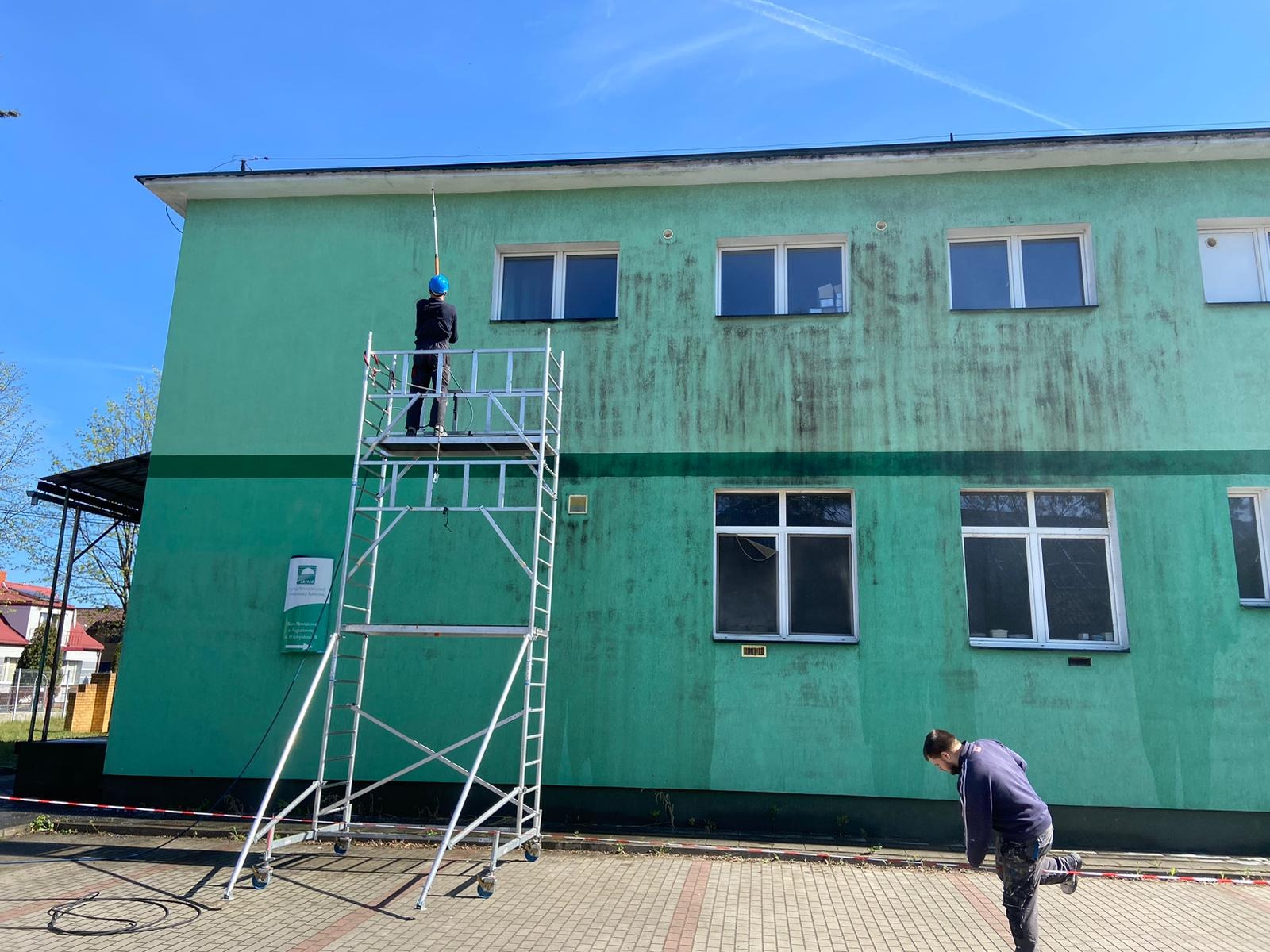 Dwóch mężczyzn przy zielonej elewacji budynku. Jeden na rusztowaniu, drugi na ziemi. Widoczne ślady zabrudzeń na ścianie, sugerujące proces czyszczenia.