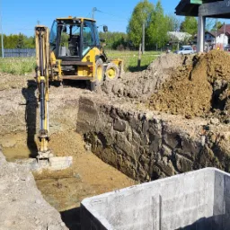 Żółta koparko-ładowarka podczas prac ziemnych, wykop pod fundamenty, widoczny betonowy szalunek oraz hałda ziemi, w tle budynki mieszkalne i zieleń.