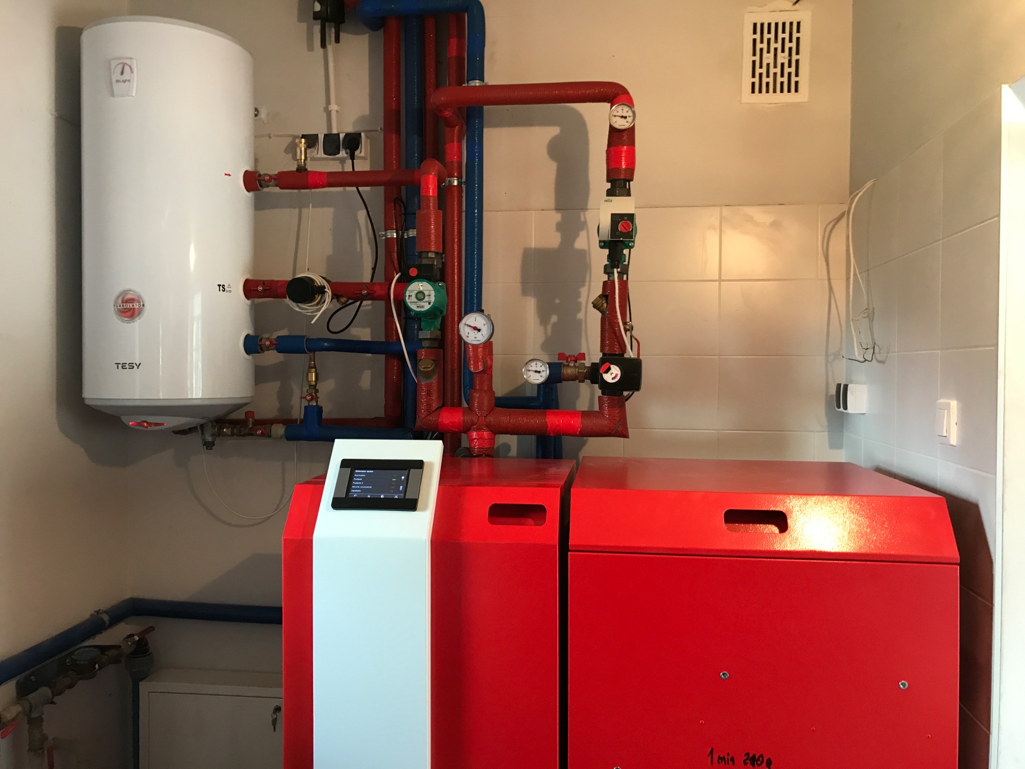 Kotłownia z czerwonym kotłem, białym zasobnikiem ciepłej wody TESY i instalacją grzewczą z rurami w kolorach czerwonym i niebieskim.