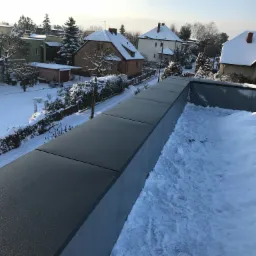 Nowoczesny, płaski dach pokryty szarymi panelami w zimowej scenerii, z widokiem na zasypane śniegiem domy i drzewa.