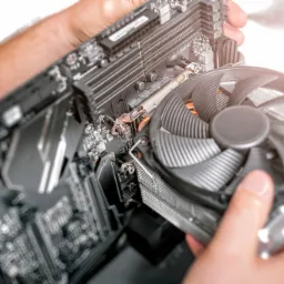 Montaż chłodzenia procesora na płycie głównej komputera, widoczne ręce technika, płyta główna z elementami, wentylator chłodzący z radiatorem, jasne tło.