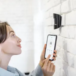 Kobieta obsługuje aplikację alarmową na smartfonie, stojąc obok czujnika na białej ceglanej ścianie.