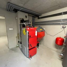 INSTAL - MIX Kamil Jezierski - Nowoczesny, czerwono-szary piec na paliwo stałe marki Defro w surowym pomieszczeniu kotłowni z widocznymi rurami instalacji grzewczej pod sufitem i czerwonym zbiornikiem wyrównawczym. Na podłodze...