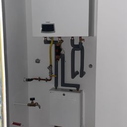 INSTAL - MIX Kamil Jezierski - Biały piec gazowy marki Termet zamontowany na ścianie, z widocznymi rurami gazowymi i hydraulicznymi, oraz skrzynką rozdzielczą poniżej, w surowym wnętrzu z betonową podłogą.