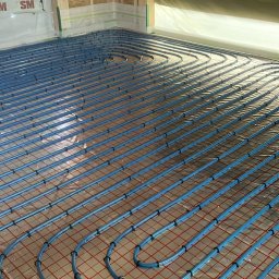 INSTAL - MIX Kamil Jezierski - Niebieskie rury ogrzewania podłogowego ułożone na czerwonej siatce w surowym wnętrzu, widoczne elementy konstrukcyjne ścian i folia ochronna.