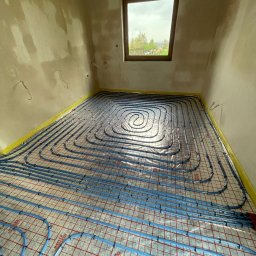 INSTAL - MIX Kamil Jezierski - Niebieskie rury ogrzewania podłogowego ułożone spiralnie na siatce w surowym pomieszczeniu z oknem, zabezpieczone taśmą przy ścianach.