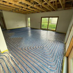 INSTAL - MIX Kamil Jezierski - Niebieskie rury ogrzewania podłogowego ułożone na folii z czerwoną siatką w surowym wnętrzu z drewnianą konstrukcją stropu i oknami z widokiem na zieleń.