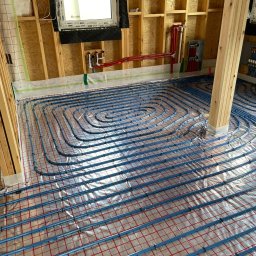 INSTAL - MIX Kamil Jezierski - Instalacja ogrzewania podłogowego w nowym budynku, widoczne niebieskie rury układane spiralnie na srebrnej folii z czerwoną siatką, rozdzielacz na ścianie, surowa konstrukcja drewniana budynku.