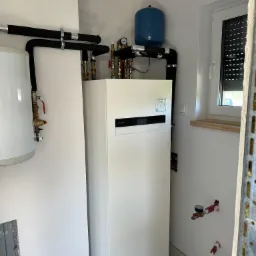 Nowoczesna, biała pompa ciepła Panasonic Aquarea Smart Cloud, z podłączonym zbiornikiem wyrównawczym w kolorze niebieskim i orurowaniem izolowanym czarną pianką, zamontowana w pomieszczeniu z oknem...