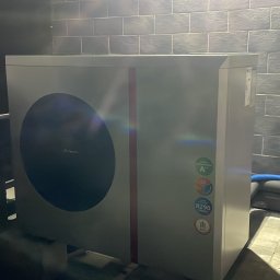 INSTAL - MIX Kamil Jezierski - Biała pompa ciepła powietrze-woda zamontowana w pomieszczeniu z szarymi płytkami na ścianie, widoczne oznaczenia klasy energetycznej A+ i czynnika chłodniczego R290.