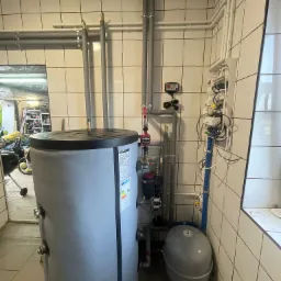 Szary bojler z podłączonymi rurami i osprzętem w kotłowni wyłożonej kafelkami, widoczne elementy instalacji i zawory.
