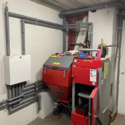 Czerwono-szary piec na paliwo stałe Defro z otwartymi drzwiczkami, zainstalowany w kotłowni z widoczną instalacją grzewczą i białą skrzynką sterowniczą na ścianie, rury zabezpieczone izolacją,...