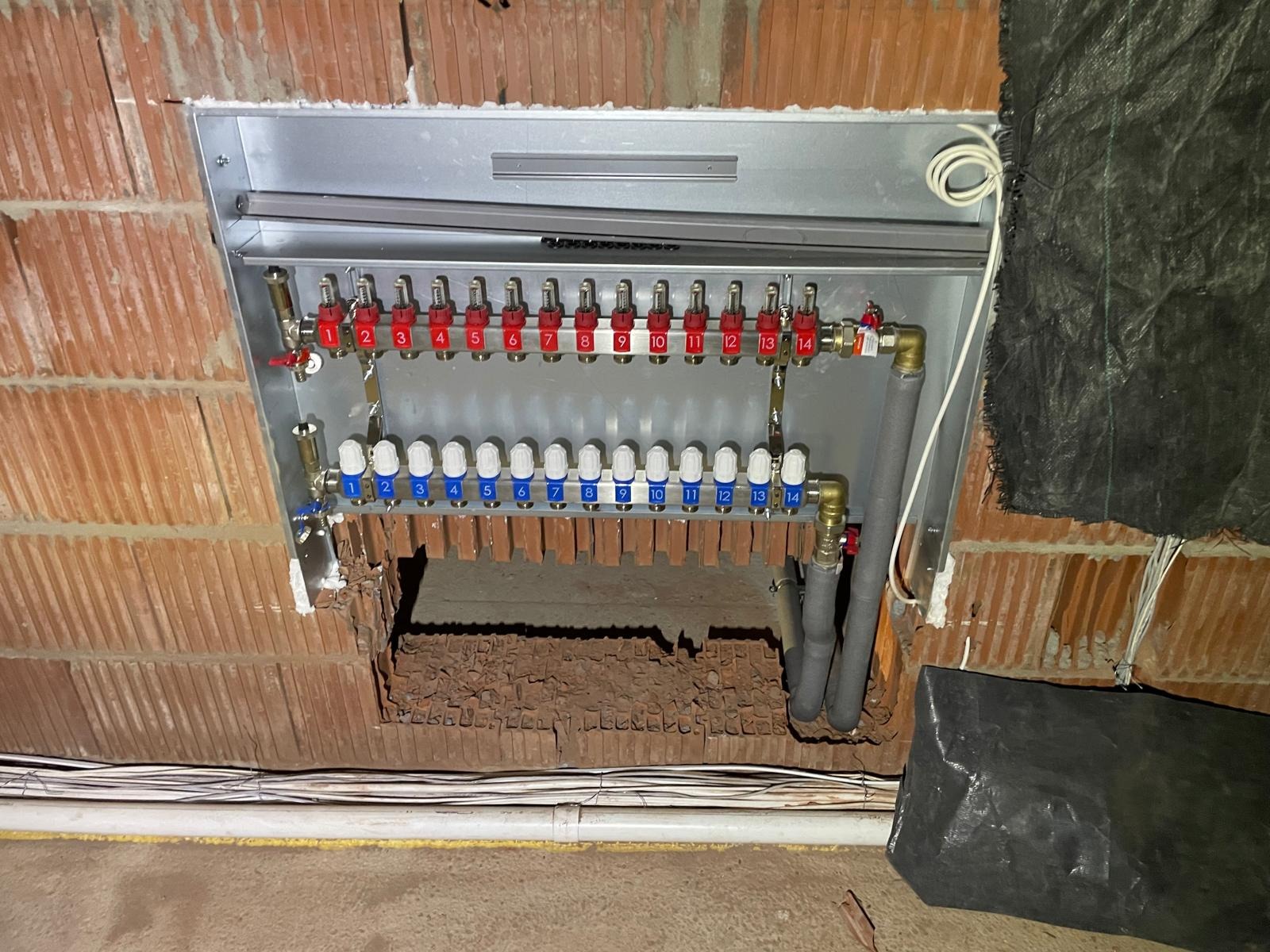 Rozdzielacz ogrzewania podłogowego z numerowanymi zaworami w metalowej szafce, zamontowany w surowej ścianie z cegły, widoczne rury doprowadzające i instalacja elektryczna.